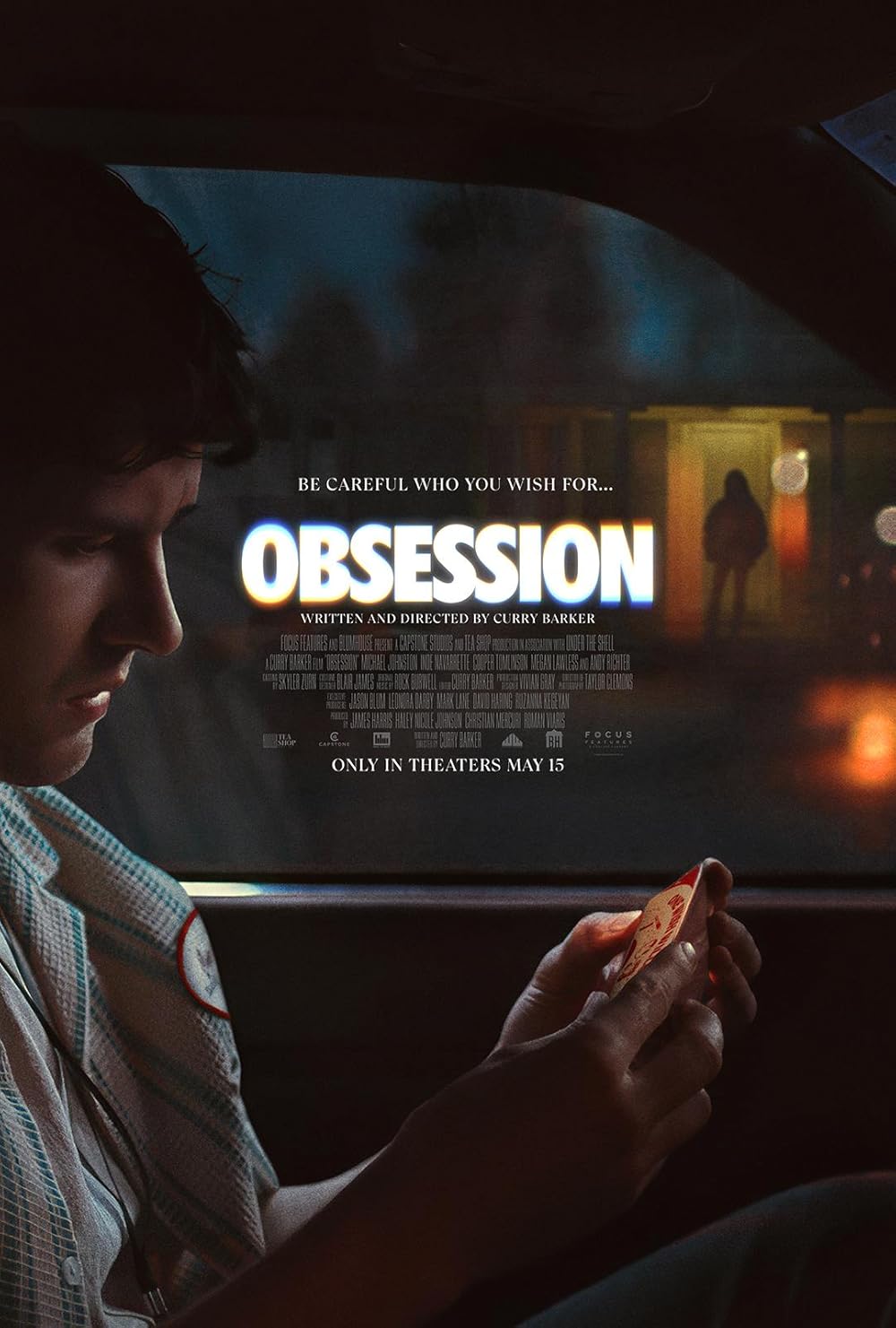 An actor is sitting in the car, looking at the phone. In the background, the house door is open with someone there.
